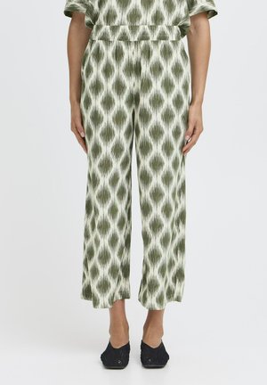 Kvinde iført grønne og hvide mønstrede vidde-bukser, der er kortede, med elastisk talje og sorte vævede sko, stående mod en ensfarvet baggrund.