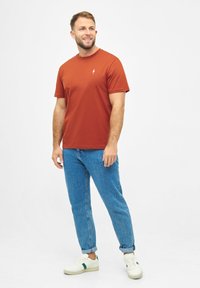 Roestkleurig t-shirt met korte mouwen en een klein wit logo, gecombineerd met blauwe spijkerbroeken en witte sneakers met groene accenten.