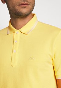 Keltainen polo-paita, jossa on teksturoitua kangasta, kaulus, kolme nappia ja kauluksessa vaaleanpunaisia raitoja. Kankaassa brodeerattu logo rinnassa.