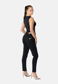 Nainen, jolla on pitkä ruskea poninhäntä, pukeutuneena mustaan hihattomaan toppiin, mustiin farkkuihin ja beigenvärisiin korkokenkiin, seisoo käsivarret lanteilla ja katsoo ylitse olkapään.