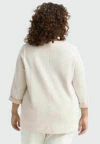 Persona con capelli ricci castani indossa una maglia a maniche lunghe beige chiaro a costine con polsini arrotolati, in piedi di spalle su uno sfondo chiaro.