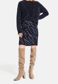 Pull en maille bleu marine, jupe à motifs avec des rayures bleues et rouille, et bottes en suède beige hautes avec des talons carrés et des zips latéraux.