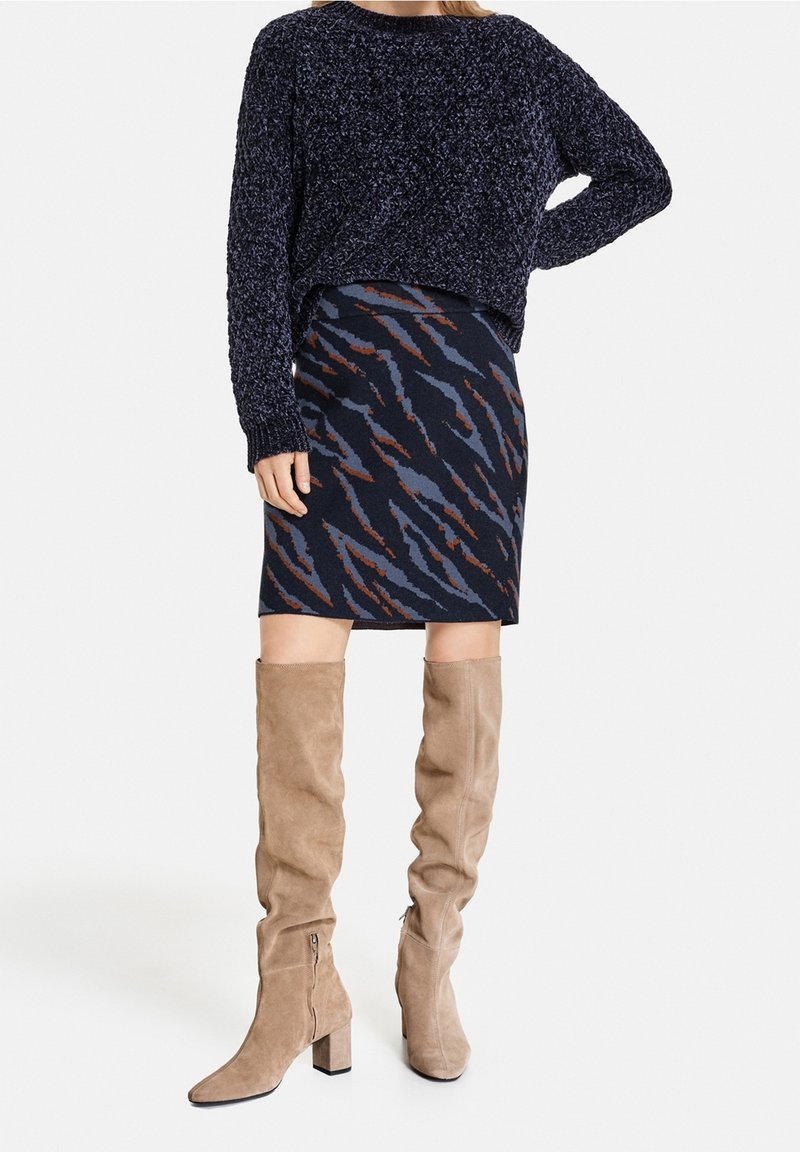 Pull en maille bleu marine, jupe à motifs avec des rayures bleues et rouille, et bottes en suède beige hautes avec des talons carrés et des zips latéraux.