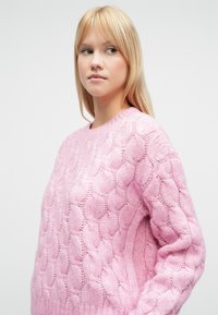 Suéter de punto rosa con un patrón texturizado de cables elevados. Tiene un cuello redondo y mangas largas, que le otorgan un aspecto acogedor.