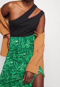 Musta yhden olkapään toppi leikkauksella, yhdistetty vihreään marmoroituun kuviolliseen hameeseen, kerroksittain kamelinvärisen bleiserin kanssa. Rakennepintainen kangas.