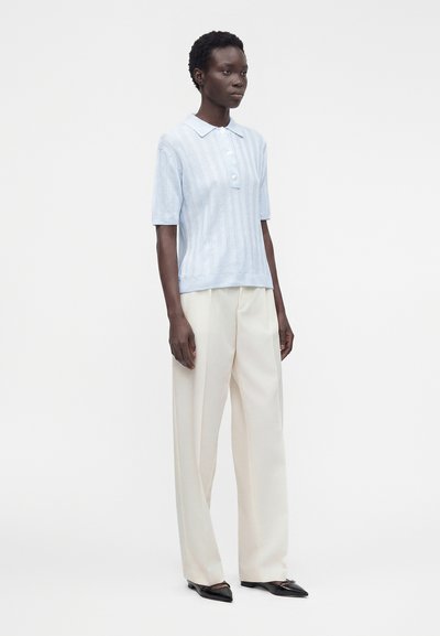 Femme debout portant une chemise bleu clair à manches courtes avec col, un pantalon crème à jambes larges et des ballerines noires pointues sur un fond uni.