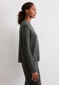Dunkelgrauer Strickpullover mit strukturiertem Muster, überschnittenen Schultern und langen Ärmeln. Trägt dazu glänzende schwarze Hosen, seitlich dargestellt.