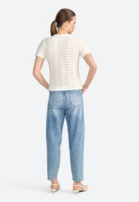 Kortærmet hvid sweater med en bølgemønster, parret med lyseblå højtaljede jeans og beige hæle.