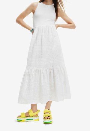 Femme portant une robe blanche à volants sans manches avec des détails ajourés, ainsi que des sandales compensées jaunes avec des accents verts et blancs.