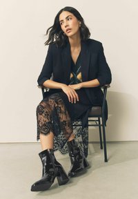 Czarne lakierowane botki za kostkę ze kwadratowym noskiem, wysokim słupkowym obcasem i bocznym zamkiem, zestawione z czarną koronkową spódnicą i marynarką.