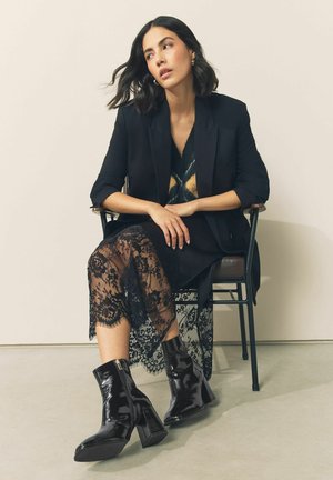 Czarne lakierowane botki za kostkę ze kwadratowym noskiem, wysokim słupkowym obcasem i bocznym zamkiem, zestawione z czarną koronkową spódnicą i marynarką.