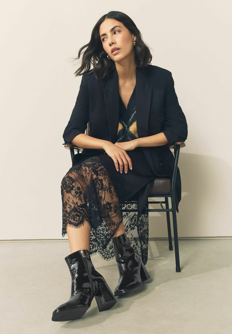 Czarne lakierowane botki za kostkę ze kwadratowym noskiem, wysokim słupkowym obcasem i bocznym zamkiem, zestawione z czarną koronkową spódnicą i marynarką.