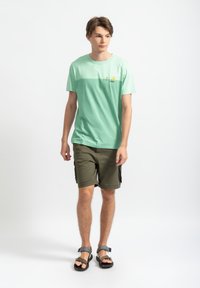 Lichtgroene katoenen T-shirt met een gradient ontwerp, voorzien van een kleine bootafdruk. Gecombineerd met olijfgroene cargoshorts en grijze sandalen.