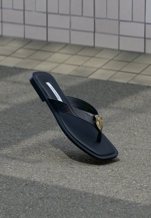 Μαύρο σανδάλι Steve Madden με πάτο τύπου flip-flop, με καπιτονέ λουράκι και διακοσμητικό σε σχήμα χρυσής καρδιάς, ακουμπημένο σε άσφαλτο κοντά σε πλακωμένο τοίχο.