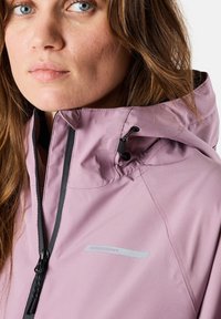 Ljust rosa jacka med huva, med svart dragkedja och snören, samt en diskret logotyp. Slät textur med minimalistisk design.