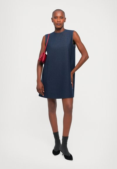 Jūras zila bezatverama kleita ar brīvu griezumu, ar sānu kabatām, apvienota ar tumši pelēkām zeķēm un melniem papēžu apaviem, turētāja sarkanā somā.