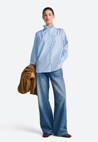 Striped blue cotton shirt with a ruffled collar, long sleeves, and single chest pocket, paired with wide-leg denim jeans and a brown jacket.