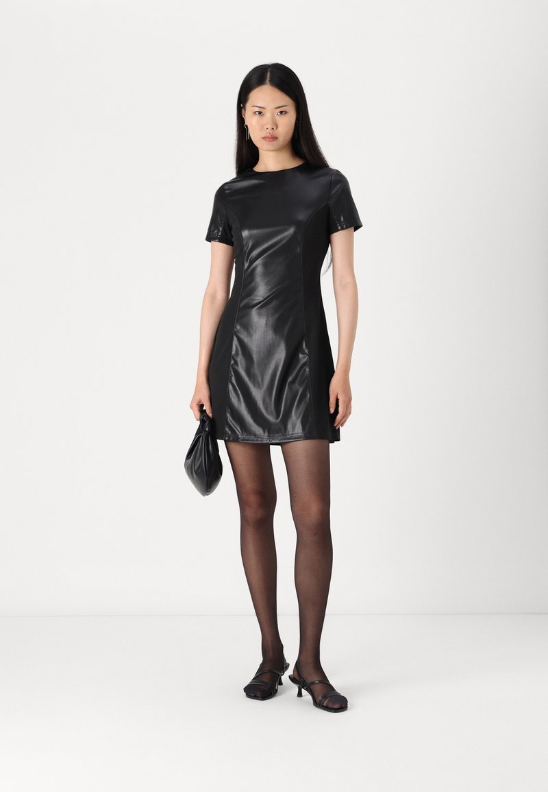 Kurzes schwarzes synthetisches Kleid mit einer schmalen Passform, kurzen Ärmeln und glänzender Oberfläche. Getragen mit durchsichtigen schwarzen Strumpfhosen und schwarzen Schuhen.