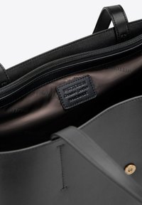 Sac fourre-tout en cuir noir avec une fermeture éclair sur le dessus, comportant une poche intérieure et une étiquette en cuir. L'intérieur est en tissu lisse de couleur sombre. Quincaillerie en métal doré visible.