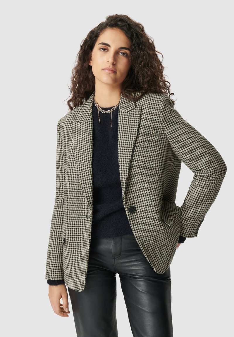 Blazer a scacchi in tessuto nero e beige, design strutturato con rever a punta, chiusura con un bottone e due tasche frontali.