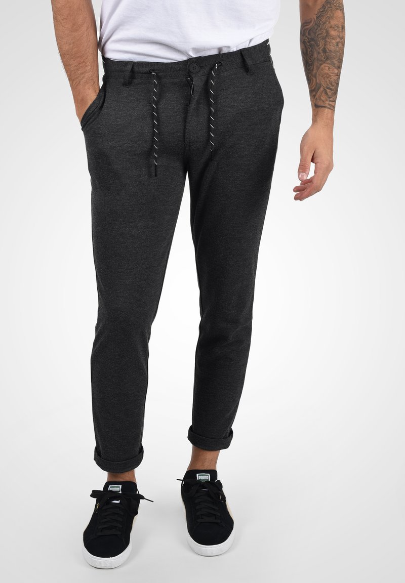 Pantalones jogger gris oscuro con cintura ajustable, dobladillos acampanados, bolsillos laterales y una textura suave y lisa. Combinados con zapatillas negras.