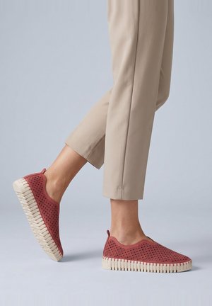Person trägt beige Hose und rote Slipper mit strukturierten Sohlen, ein Fuß angehoben, vor einem einfarbigen hellen Hintergrund.