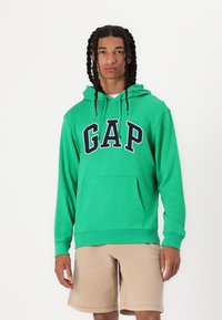 Felpa verde con cappuccio con la scritta "GAP" in blu navy. Presenta una tasca frontale e polsini a coste. Indossata con pantaloni corti beige.