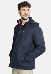 Chaqueta aislante de color azul marino con cierre frontal de cremallera, dos bolsillos laterales y capucha. Presenta un patrón texturizado en el forro interior.