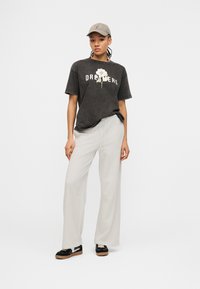 Γυναίκα με γκρι καπέλο, μαύρο t-shirt "Dreamers" με γραφικό λουλούδι, ανοιχτό μπεζ παντελόνι φαρδιάς γραμμής και μαύρα αθλητικά παπούτσια με λευκές λεπτομέρειες.