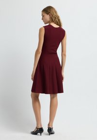 Robe sans manches couleur bourgogne avec un corsage ajusté et une jupe évasée, présentant une texture à rainures verticales et des lignes épurées, assortie de chaussures noires.