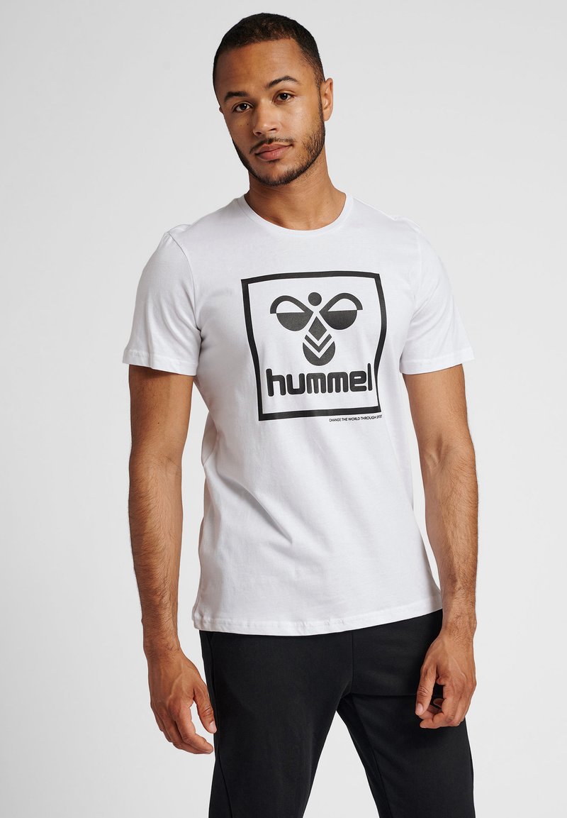 Camiseta blanca de algodón con un logo cuadrado negro que presenta una abeja estilizada y el texto "hummel!" debajo. Diseño de manga corta y cuello redondo.