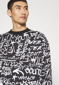 Felpa nera con testo e motivi in stile graffiti bianchi. Collo rotondo, maniche lunghe, materiale in cotone con superficie testurizzata.