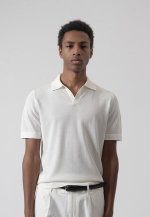 Joven con cabello corto y rizado, vestido con una camisa polo blanca de manga corta y pantalones blancos con cinturón negro, posando frente a un fondo gris liso.