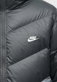 Mustmust musta jope, millel on diagonaalne tikandi disain, lukuga sulgemine ja valge Nike logo rinnal. Sile tekstuur.