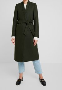 Cappotto avvolgente verde oliva realizzato in tessuto strutturato, con vita cinturata, profondo collo e design di lunghezza media, abbinato a jeans azzurri.