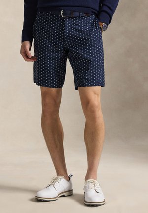 Άνδρας με μπλε σκούρο σορτς με σχέδια, λευκά παπούτσια του γκολφ, μπλε σκούρο πουλόβερ και καφέ ρολόι, στέκεται σε ουδέτερο φόντο.