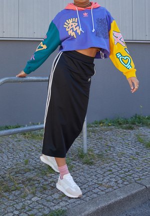 Osoba w kolorowej krótko ściętej bluzie z kapturem i czarnej spódnicy Adidas opiera się o metalową poręcz na brukowanym chodniku na zewnątrz.