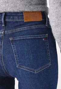 Jeans de denim azul oscuro con un primer plano de la parte trasera que muestra una etiqueta de cuero con la inscripción "TOMMY HILFIGER" y costuras en contraste.