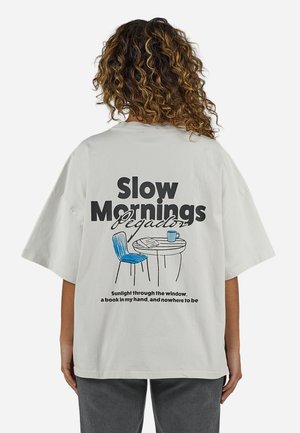 Göndör hajú személy világosszürke ingben, aki egy széket és egy asztalt mutat, amelyen egy bögre áll, a hátán pedig a "Lassú reggelek" felirat olvasható.