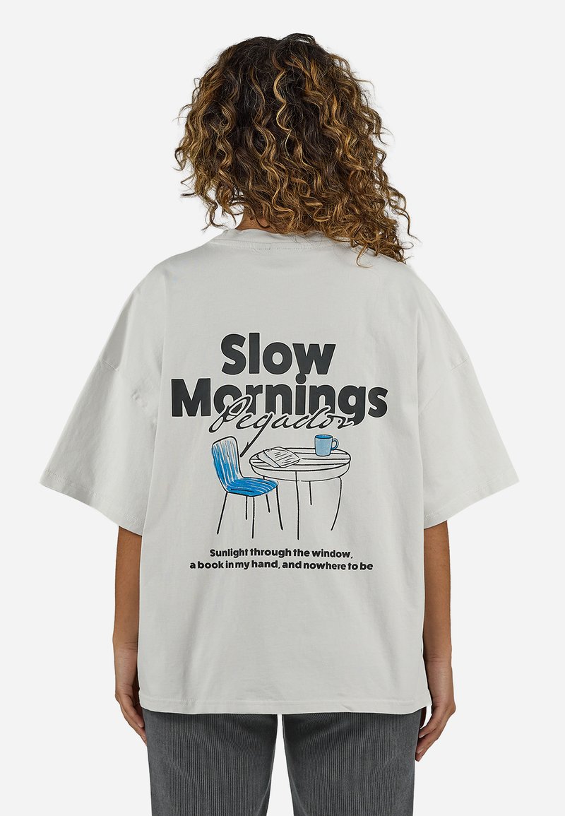 Άτομο με σγουρά μαλλιά που φορά ένα ανοιχτό γκρι πουκάμισο δείχνοντας μια καρέκλα, τραπέζι με κούπα και κείμενο "Αργά Πρωινά" στην πλάτη.