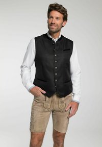 Schwarze Wolleweste mit silbernen Knöpfen, kombiniert mit beigen Lederhosen mit Blumenspitze. Ein weißes Hemd darunter sorgt für einen Kontrast.