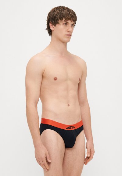 Μαύρα εσώρουχα με κοντό μήκος και με αντίθετη πορτοκαλί ζώνη που φέρει λογότυπο. Το ύφασμα φαίνεται λείο και ελαφρώς ελαστικό.