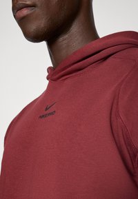 Bordeauxroter Nike Pro Hoodie mit schwarzem Logo und Text. Verfügt über einen strukturierten Stoff und eine lockere Passform mit einer auffälligen Kapuze.