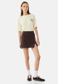 Lys gul grafisk t-shirt med et tegneserie-design, parret med en brun mini-nederdel med en sideslidse. Modellen har hvide ankelstrømper og brune sneakers på.