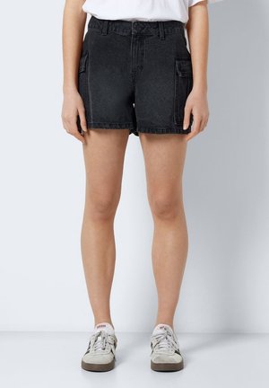 Person trägt schwarze Denim-Cargoshorts, weißes T-Shirt und weiße Adidas-Sneaker und steht vor einem schlichten Hintergrund.