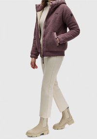 Chaqueta morada difusa con capucha y cremallera, combinada con pantalones crema y botines de ante beige. Tela texturizada y puños acanalados detallados.