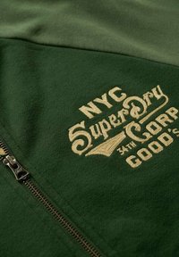 Haut zippé en tissu vert avec du texte brodé beige, portant l'inscription "NYC SuperDry 34th Corp Good's." Comprend un détail de fermeture éclair en métal.