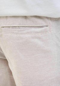 Jasnobeżowe lniane spodnie o gładkiej fakturze mają tylnią kieszeń z prostym, przeszytym brzegiem. Powyżej częściowo widoczna jest biała tkaninowa bluzka.