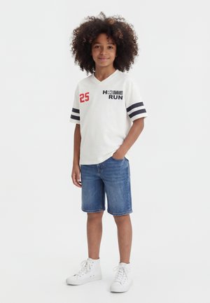 Παιδί με σγουρά μαλλιά στέκεται φορώντας ένα λευκό μπλουζάκι "Home Run" με μαύρες ρίγες, μπλε τζιν σορτς και λευκά ψηλά αθλητικά παπούτσια.