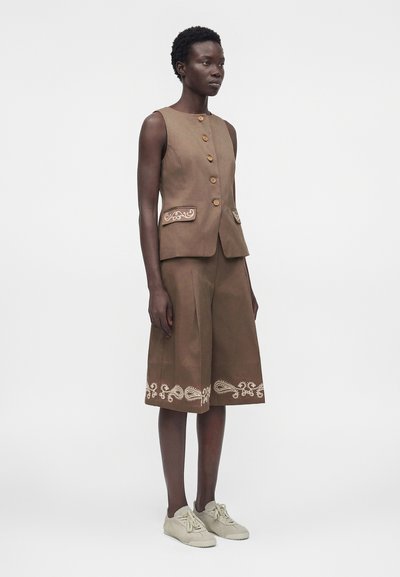 Γυναίκα που φοράει καφέ αμάνικο γιλέκο με κουμπιά και αντίστοιχα σορτς μέχρι το γόνατο με λευκή κεντητική και μπεζ αθλητικά παπούτσια, στέκεται σε λευκό φόντο.